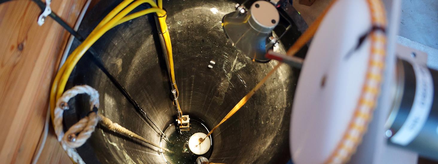 Flottørmåler, et rør med en trang åpning i bunnen er plassert ned i sjøen. Over røret henger et tannhjul med et kjede som har en flottør i den ene enden og en motvekt, et lodd, i den andre. Foto.