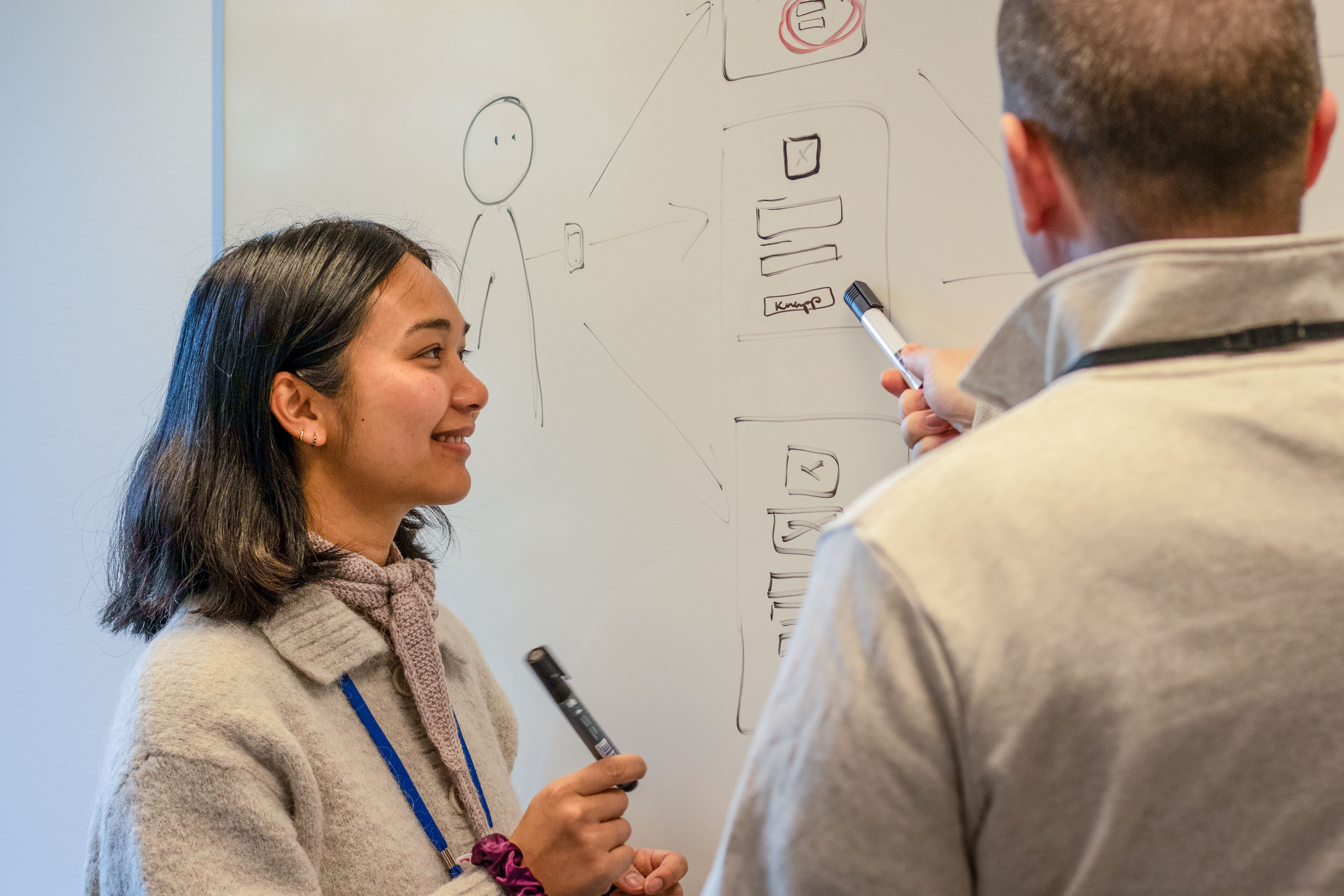 Kvinne med tusj forran et white-board med en mann i forgrunnen. 