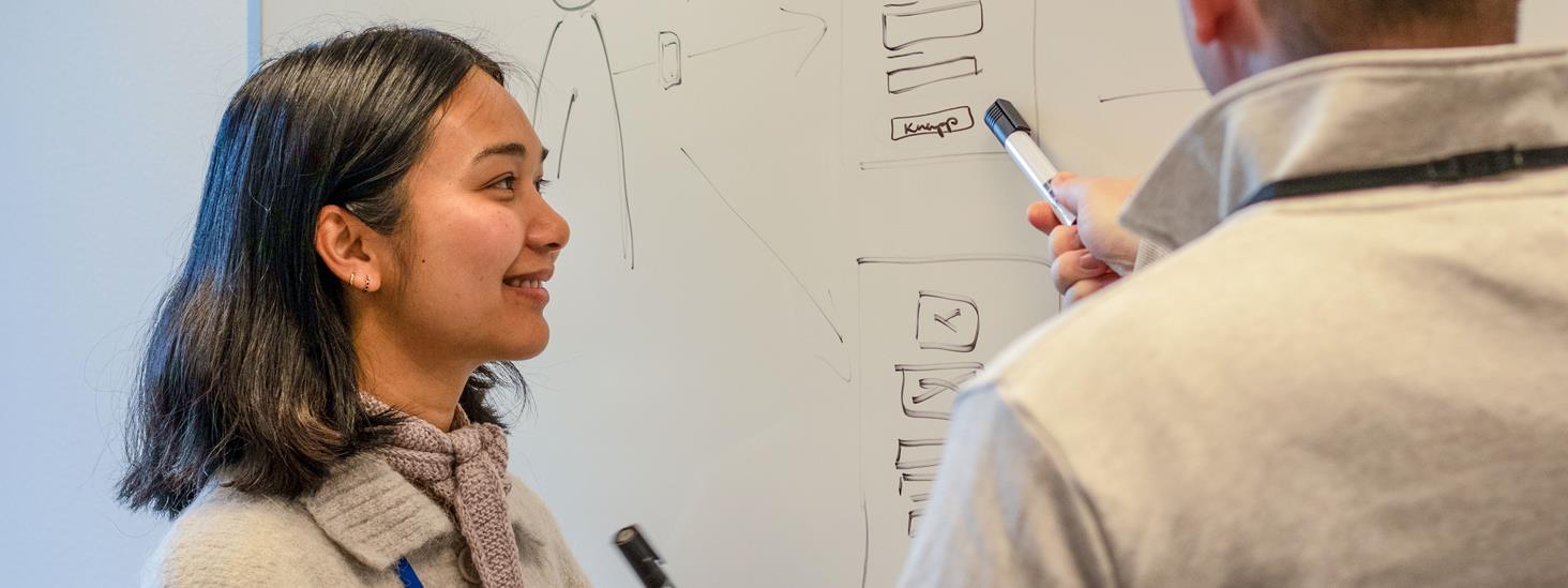 Kvinne med tusj forran et white-board med en mann i forgrunnen.