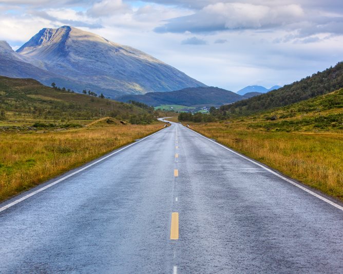 Asfaltert vei som forsvinner bak åser mot fjell.