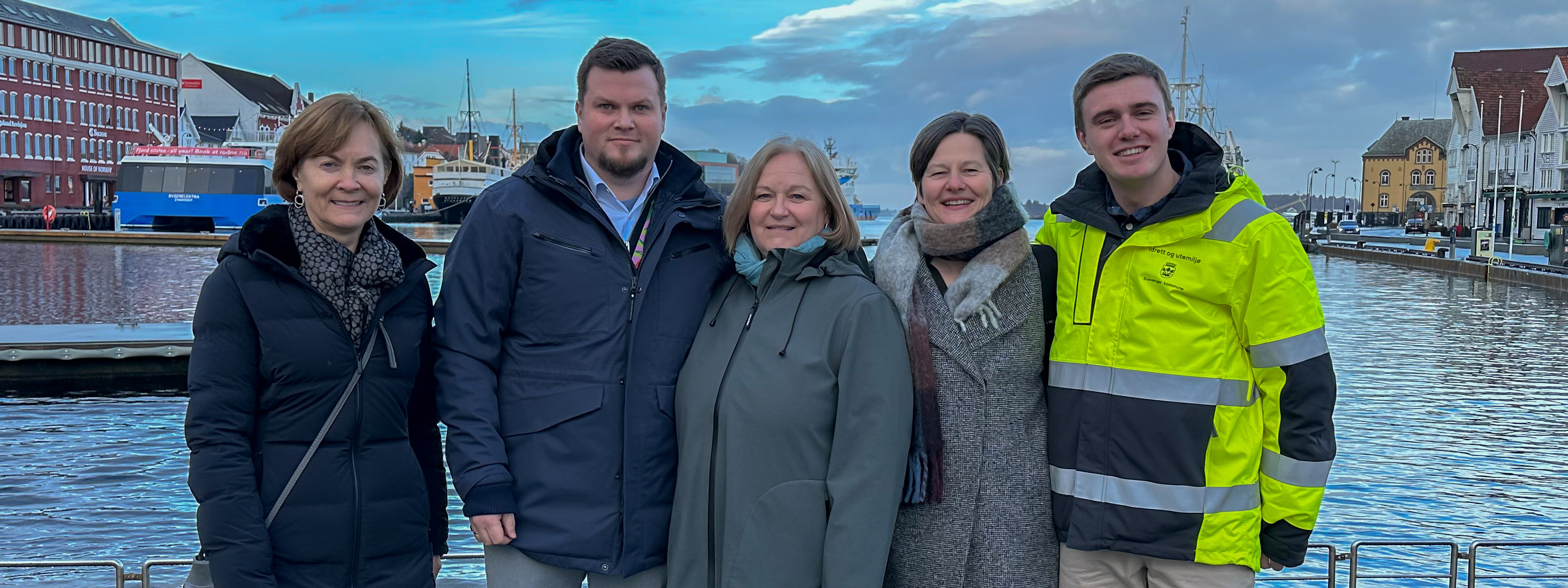 Seks voskne mennesker, tre damer og to menn, står oppstilt i Vågen i Stavanger. Alle smiler. De er kledt i vinterjakker.
