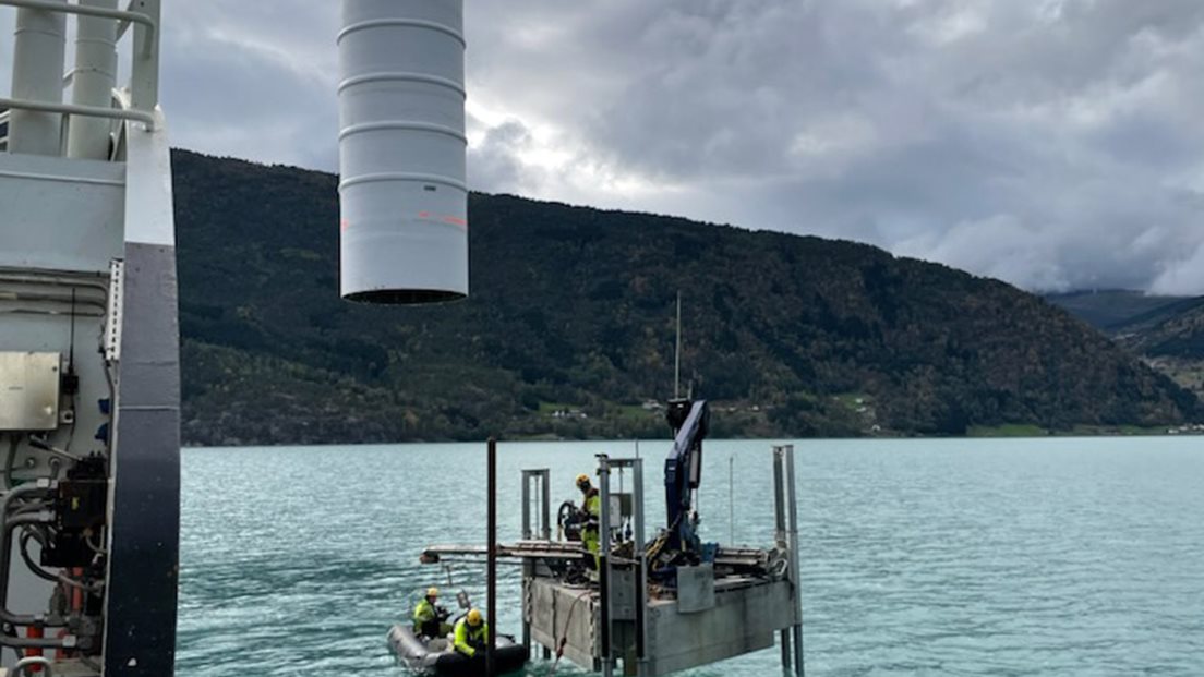 Sjømerke installeres til sjøs. En mann står på installasjonen mens to menn hjelper til fra gummibåt. Deler av skip ses i forgrunnen. Foto: Kystverket