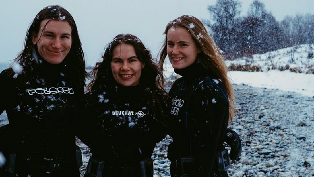 Stine Skalmerud, Juni Bjørneset og Ida Søhol, ikledd våtdrakt, står i fjæra. Det snør ute. Foto: Mauro Pau