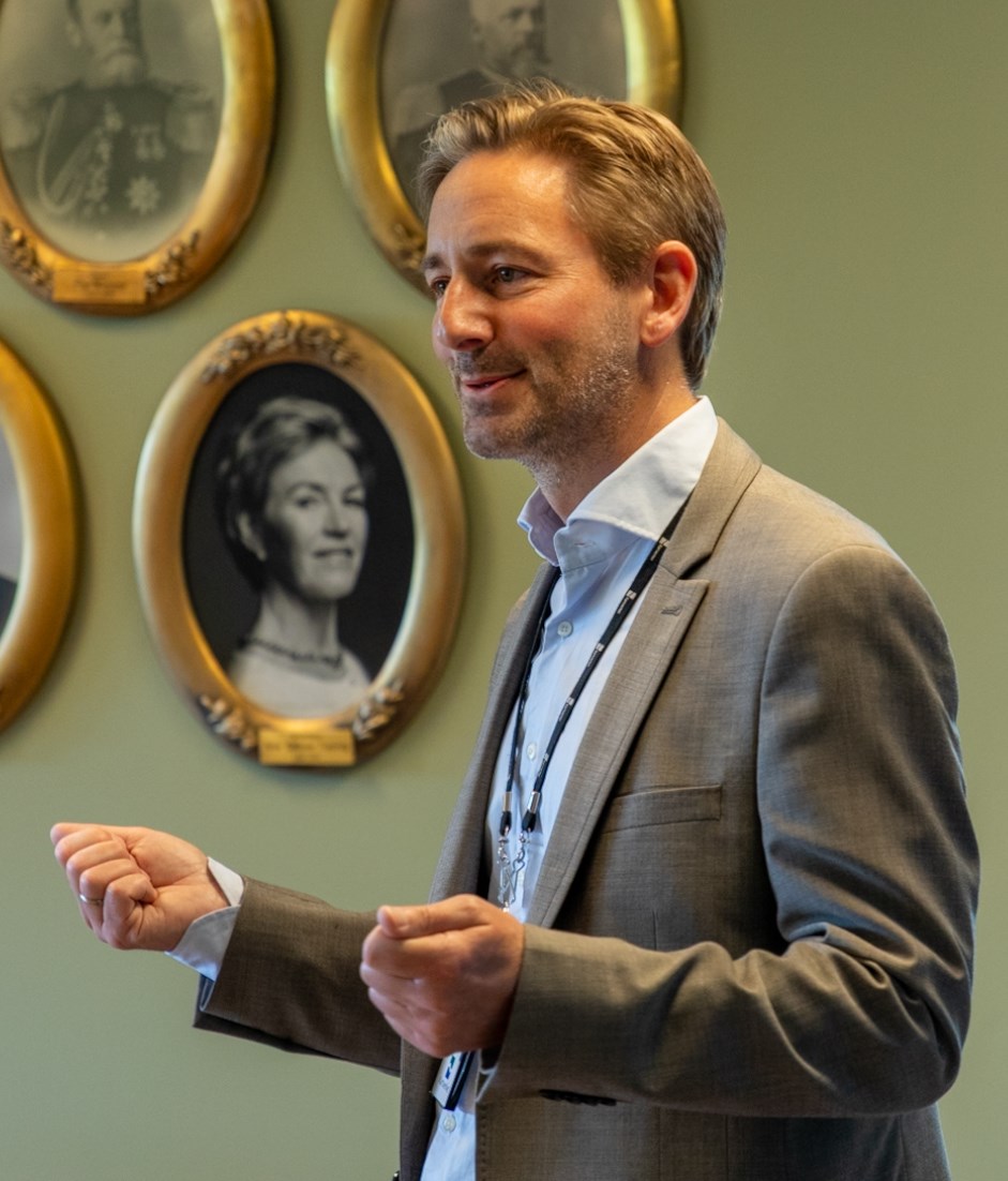 Mann i dress snakker i et rom med flere foto i bilderammer p&aring; veggen,
