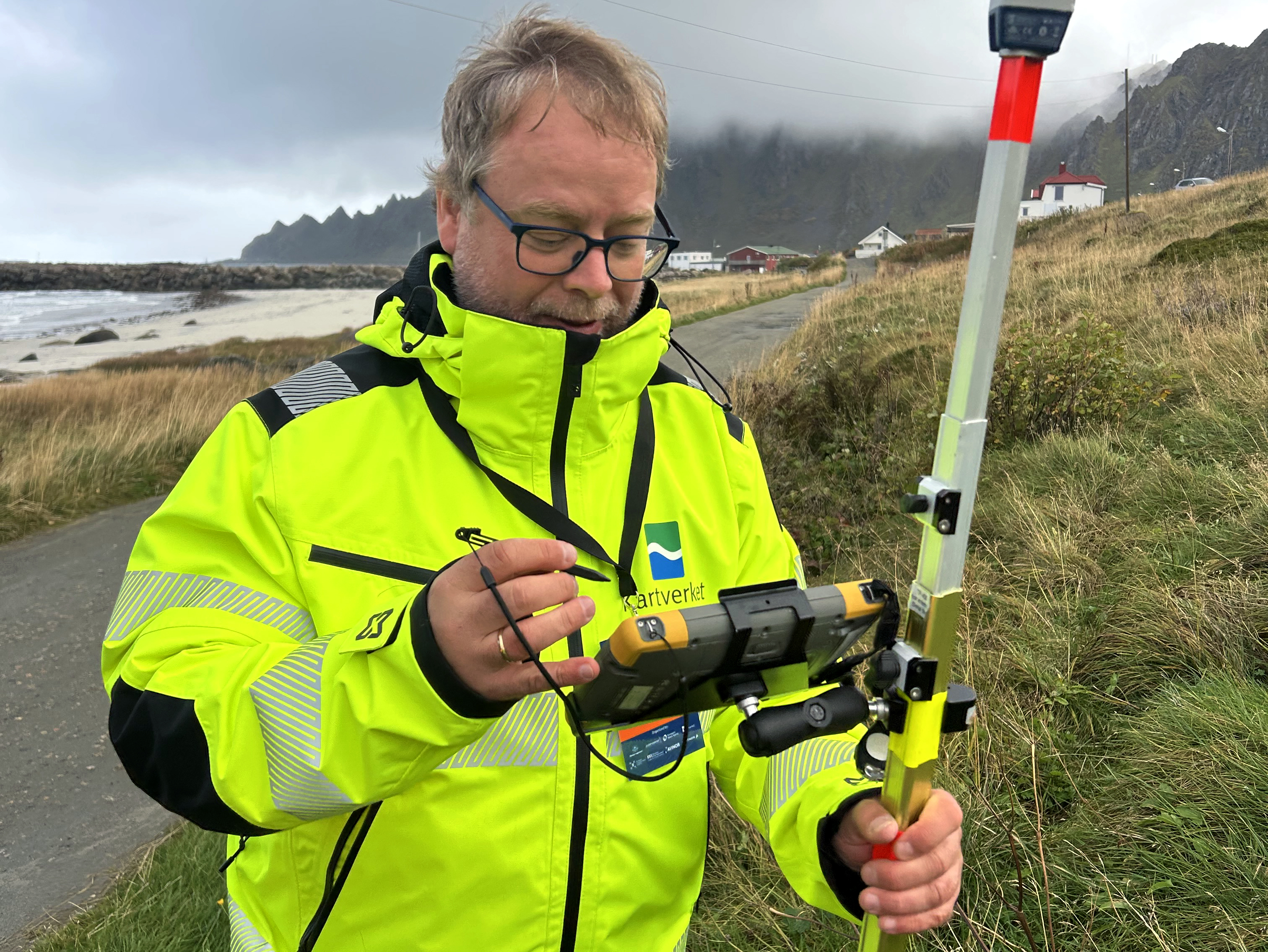 En person i gul jakke står ute ved en GPS-stasjon i et grønt landskap, med fjell i bakgrunnen.