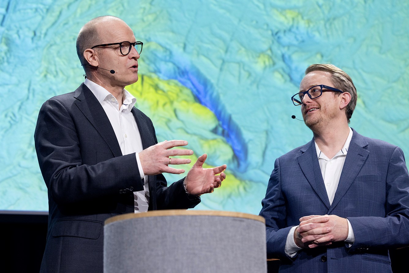 Kartverkssjef Johnny Welle og Morten Goodwin i panelsamtale p&aring; Kartverkskonferansen 2026. Terrengmodell av havbunnen vises p&aring; en skjem bak dem.