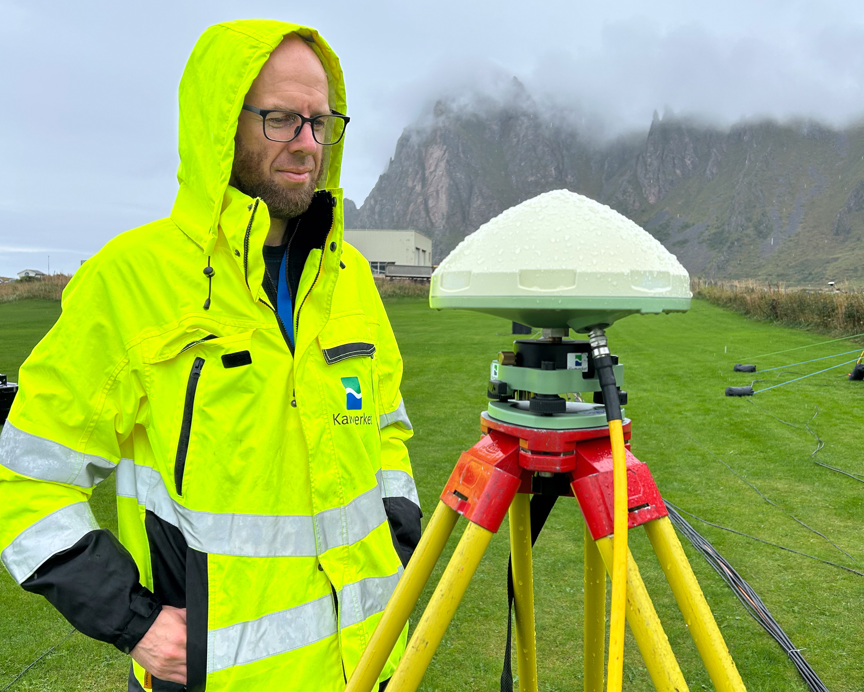En person i gul jakke står ute ved en GPS-stasjon i et grønt landskap, med fjell i bakgrunnen.