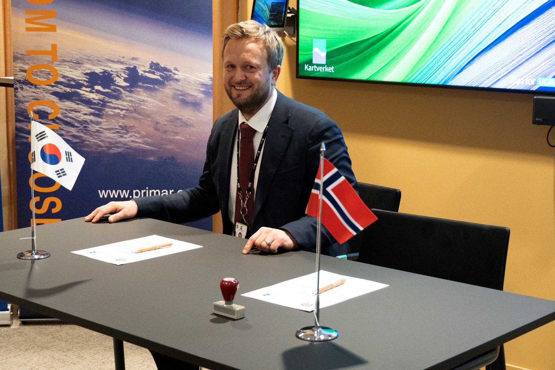 Mann idress sitter ved et bord som har to flagg st&aring;ende opp&aring;