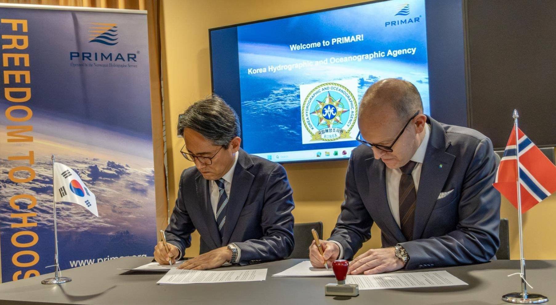 To menn signerer en avtale foran en skjerm med PRIMAR-logo. Det står et sør-koreansk og norsk flagg på bordet.