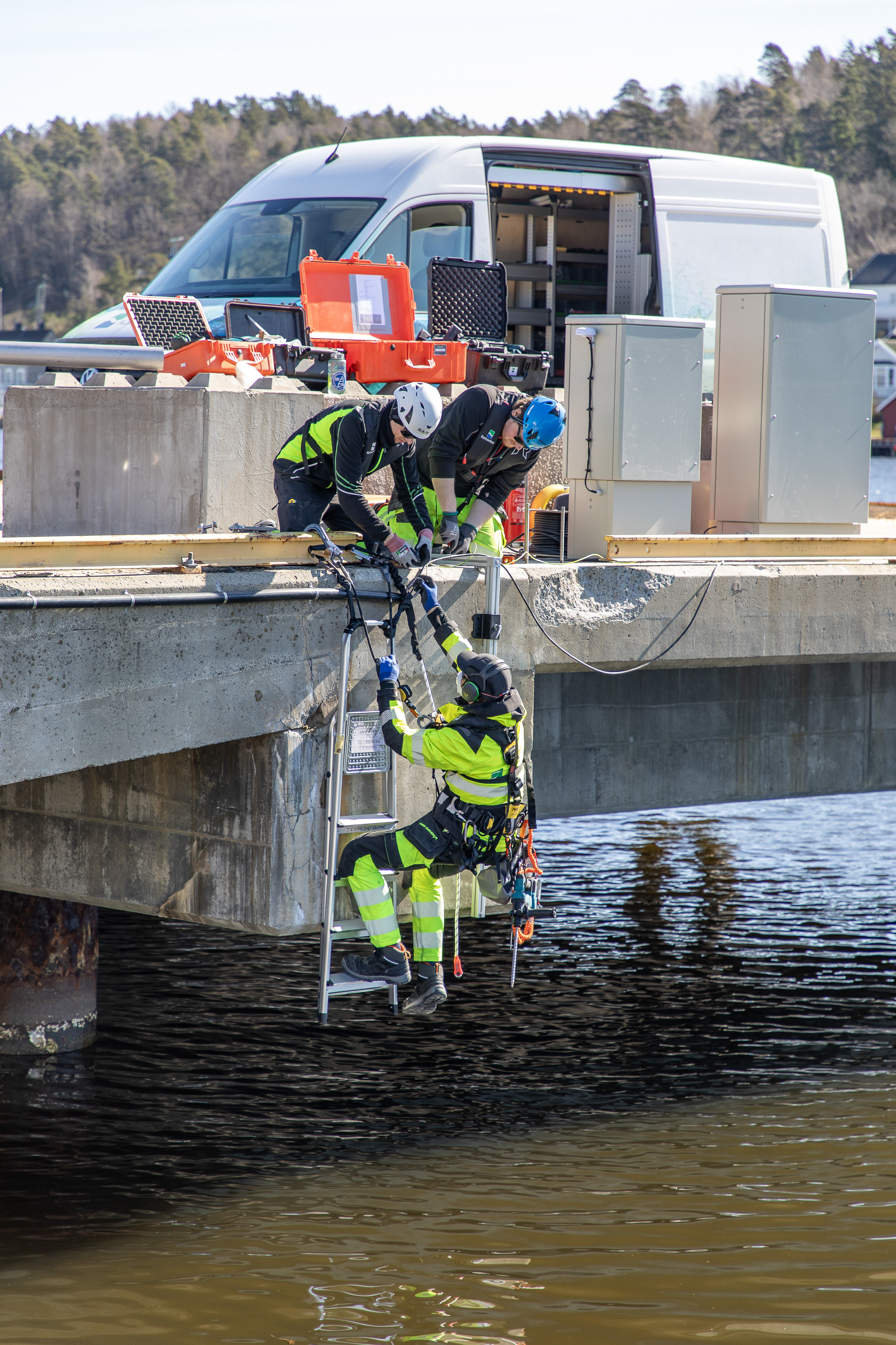 En person med klatresele st&aring;r i ei gardintrapp som er sikra med tau i kaikanten, mens to kolleger sender teknisk utstyr til ham.