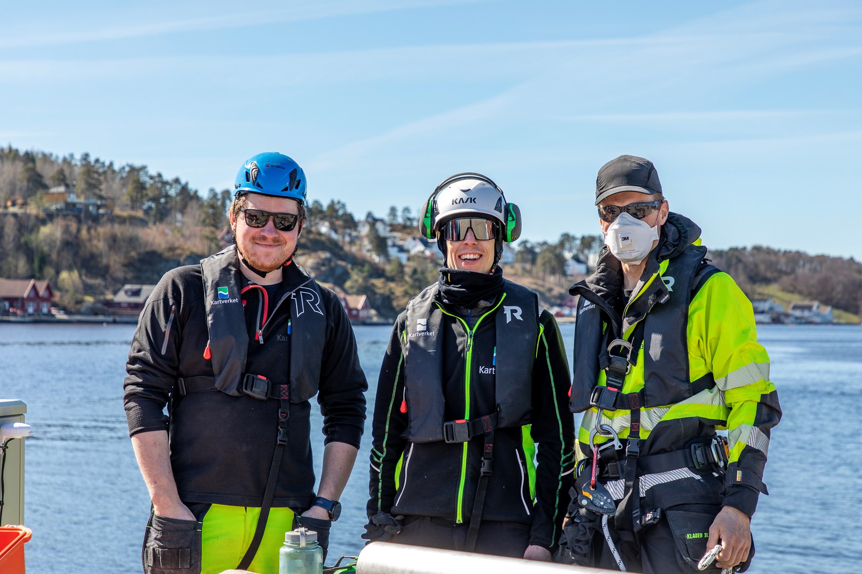 Tre menn i arbeidst&oslash;y p&aring; en kaikant smiler mot kamera. Det er fint v&aring;rv&aelig;r med stille sj&oslash; og bl&aring; himmel i bakgrunnen.