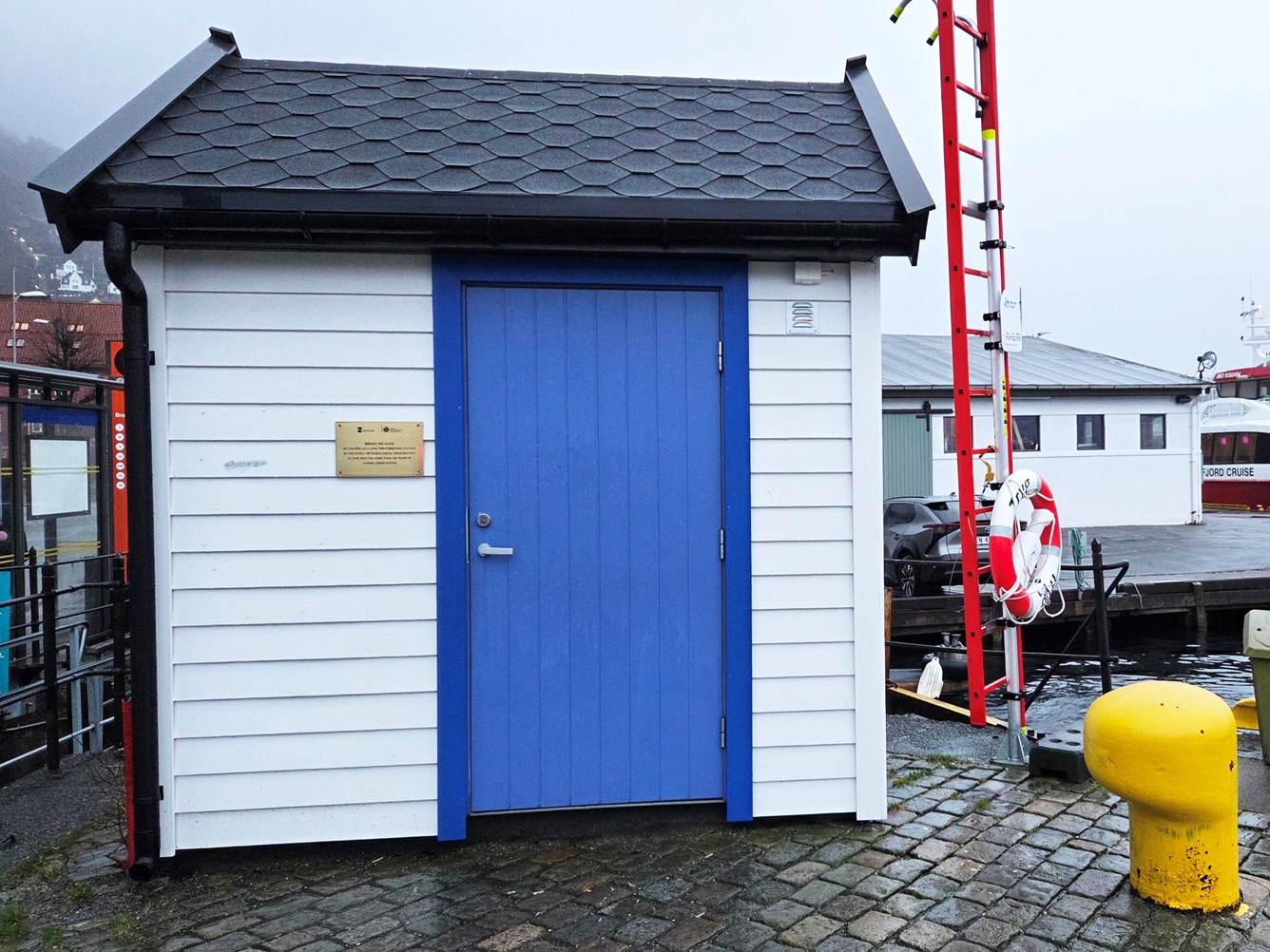 Lite hvitt bygg med knallbl&aring; d&oslash;r p&aring; ei kai i bergen. En stige med livb&oslash;ye og en pullert til h&oslash;yre i bildet.