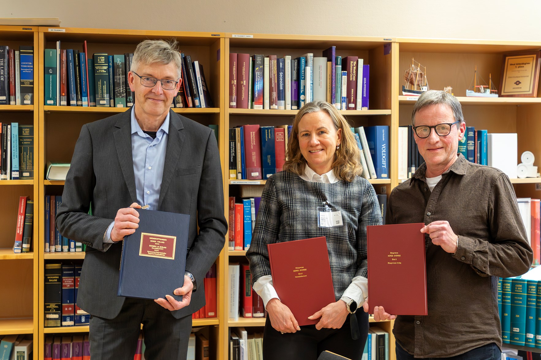 To menn og en kvinne foran en bokhylle som smiler inn i kamera. Alle holder hver sin bok i hendene.